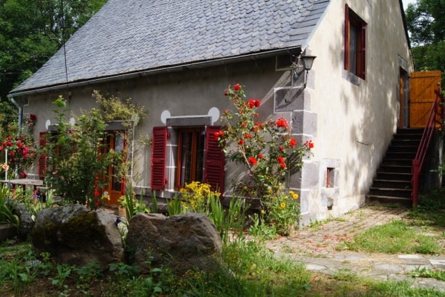 Le gîte vu de face, en été. On peut y voir des volets rouges à chaque fenêtre et une partie très fleurie.